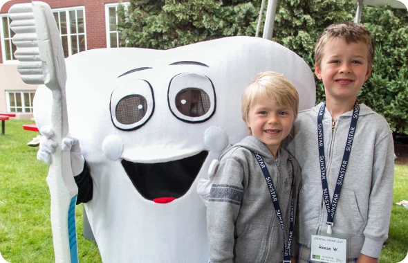 Two smiling young children stand outdoors with a tooth mascot. 