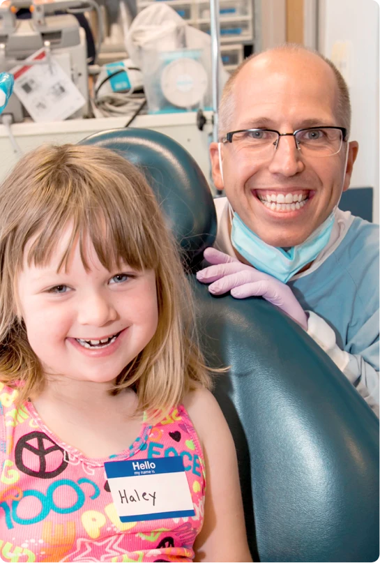 A young girl sits in an exam chair smiling while a dentist smiles behind her. 