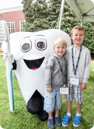 Two smiling young children stand outdoors with a tooth mascot.