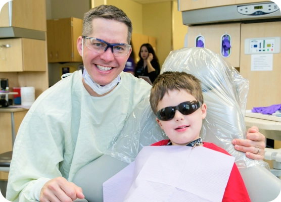 A boy wears sunglasses in an exam chair with his provider smiling next to him.