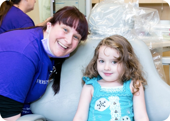 A young girl smiles in an exam chair with her provider smiling next to her.