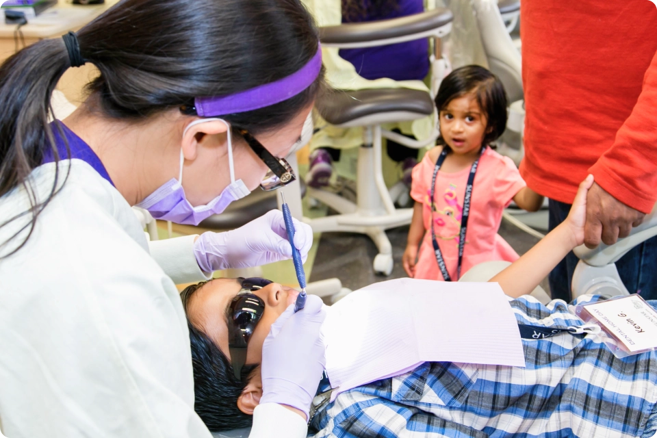 Provider working on a young patient’s teeth as a young girl stands in the background with an adult.